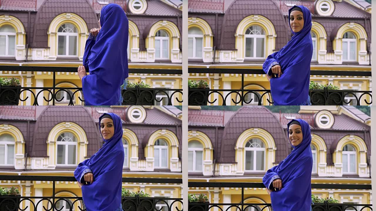 年轻美丽的穆斯林妇女戴着头巾站在阳台上，转身对着镜头微笑，背景是建筑