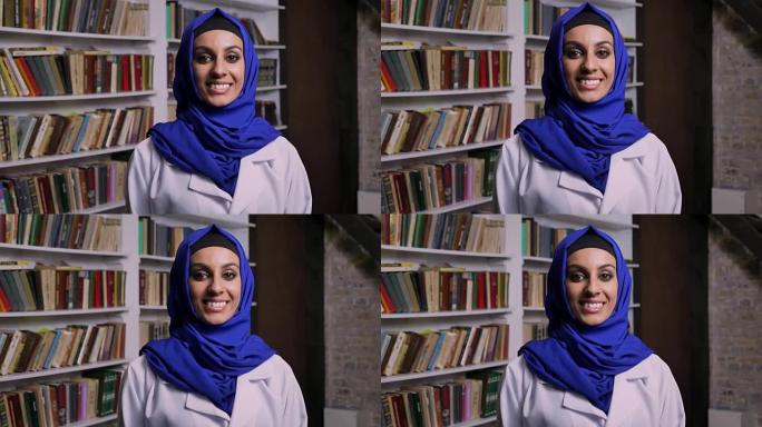 戴着头巾的年轻快乐穆斯林妇女站在图书馆，看着相机，微笑着