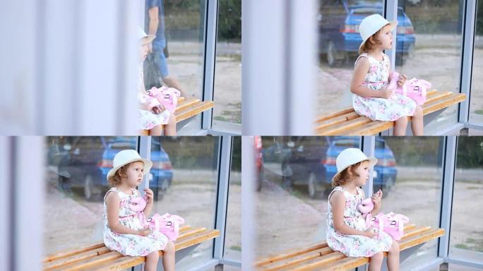 戴着帽子，背着背包的女童游客坐在公共汽车站的长椅上，等待运输以进行进一步的旅行
