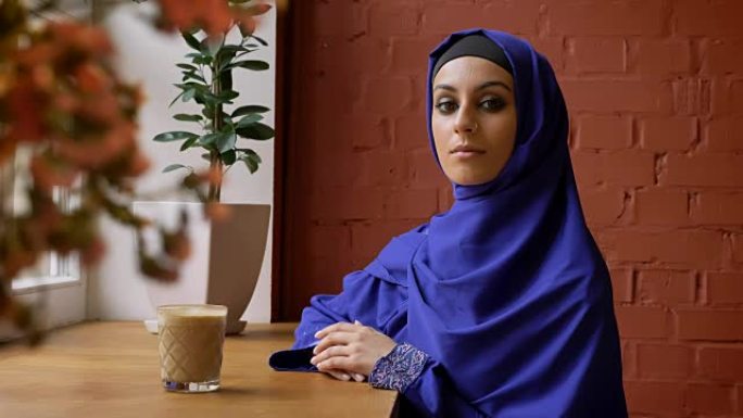 年轻的穆斯林妇女戴着头巾坐在咖啡馆里看着镜头，戴着头巾的美丽女性