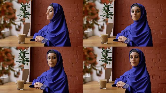 年轻的穆斯林妇女戴着头巾坐在咖啡馆里看着镜头，戴着头巾的美丽女性