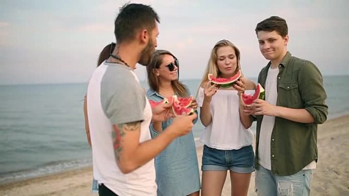 朋友站在沙滩上吃西瓜，用智能手机摄像头自拍。穿着蓝色牛仔裤短裤的年轻男女在海边聊天。健康营养，素食主