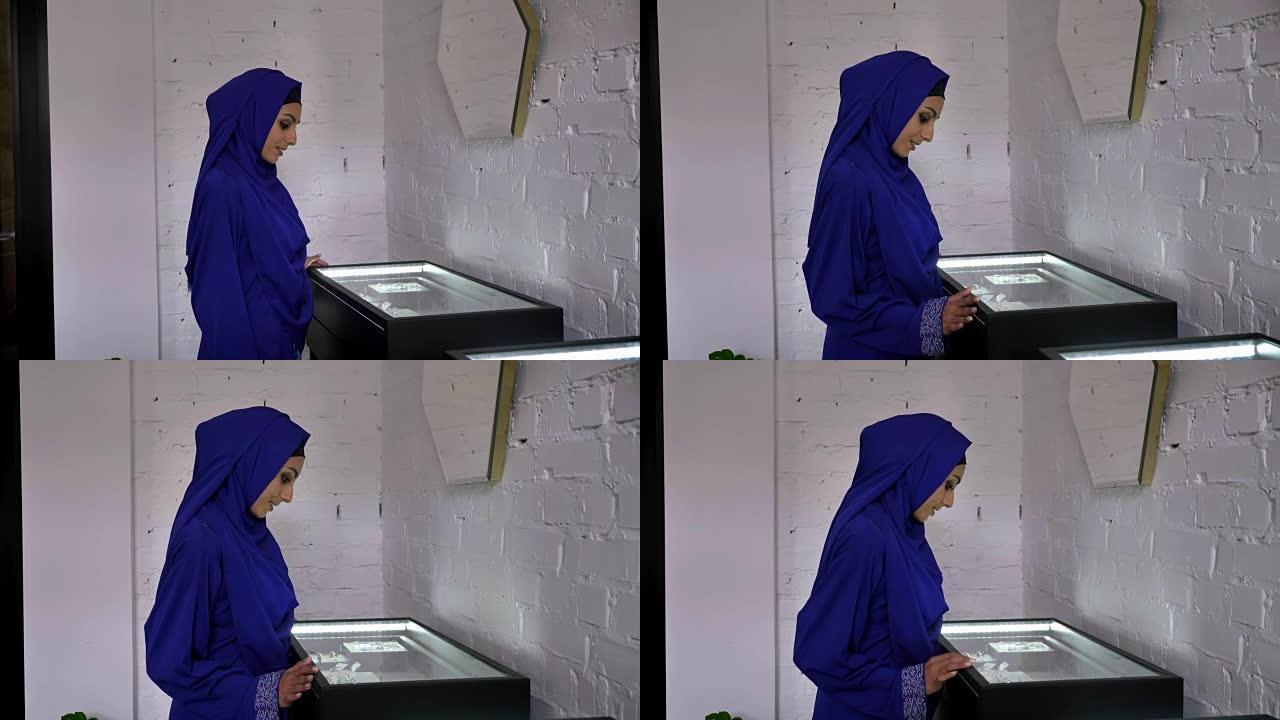戴着头巾的年轻穆斯林妇女看着珠宝店里的配饰展示，并试图决定买什么