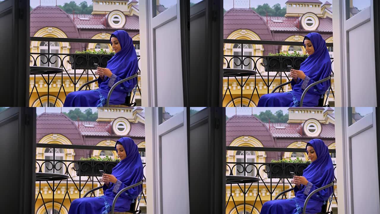 戴着头巾的年轻迷人穆斯林妇女坐在阳台的椅子上，用手机打字