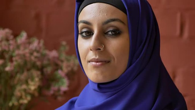 年轻美丽的穆斯林妇女戴着头巾，看着镜头，鲜花和背景墙
