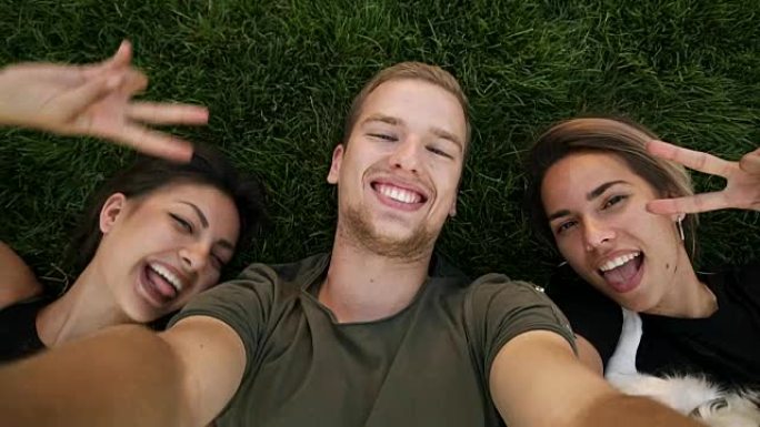 三个迷人的千禧一代朋友在公园或草坪上躺在绿色的草地上自拍。高加索男人和两个女孩一起微笑着玩得开心。从