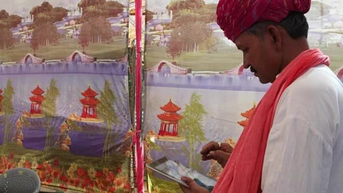 手持印度男子忙于触摸屏平板电脑，配有色彩鲜艳的帐篷背景