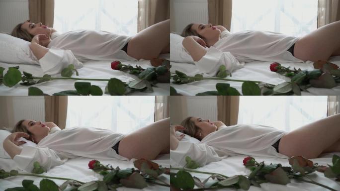 穿着透明短睡衣的甜美女士躺在床上，妆容明亮。红玫瑰在靠近女性身体的床上。慢动作。