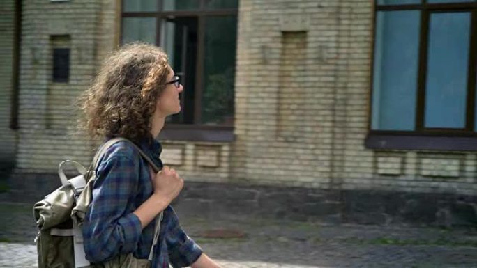 戴着眼镜的波浪形头发的聪明书呆子学生走向大学，单肩背着背包，摸着头发