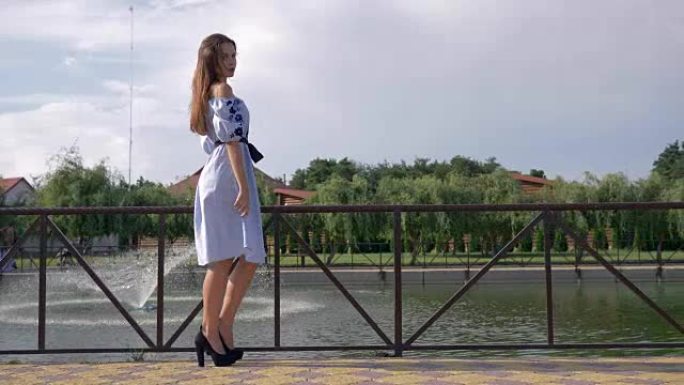 穿着裙子和高跟鞋的漂亮女孩在湖水背景下带着喷泉在户外散步