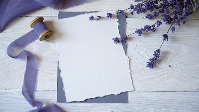 白色空白卡片，信封和缎带在粉红色和蓝色织物的背景上，淡紫色花朵在白色背景上。带有信封和空白卡的模型。