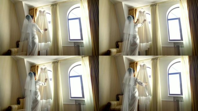 靠近窗户，在房间里，白色的婚纱挂在窗檐上。新娘，一个穿着白色便衣的漂亮女孩，穿着浴袍，正在考虑她的婚