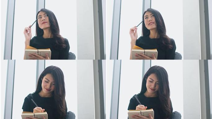 迷人的亚洲女性黑色连衣裙工作和纸上笔记坐在沙发上，窗灯从背后微笑着自信快乐