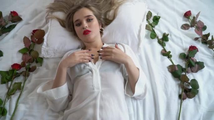 年轻女士的肖像，妆容明亮，鼻子穿孔。穿着短透明睡衣的女人躺在床上，带着红玫瑰。红玫瑰在靠近女性身体的