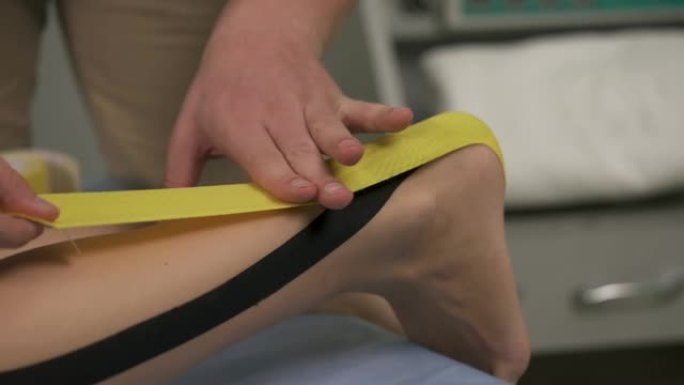 男性手的特写镜头，将黄色胶带固定在脚跟上，沿小腿拉伸并用纸背衬摩擦。物理治疗师正在运动跟腱。