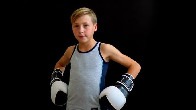 一个年轻的运动员站在黑暗的背景上。这个男孩穿着灰色t恤，双手戴着手套。黑白手套部分可见。这个男孩有金