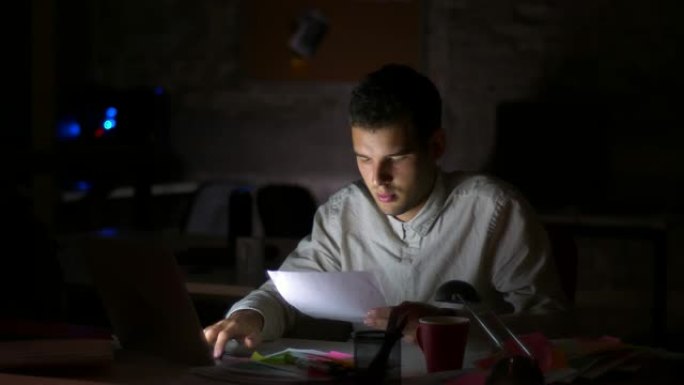 拍摄黑暗的办公室，屏幕上的光线照在他的脸上，而他正坐着看电脑和检查文件，用专注的自信的脸拿着它们，室