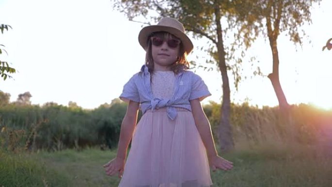 日落时分，戴着墨镜和草帽的小女孩在摄像机前跳舞