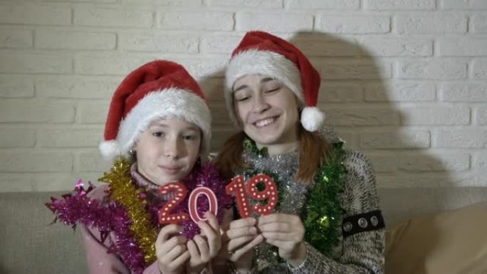 两个快乐的小女孩展示了相机的数字2019。他们坐在沙发上的圣诞老人的帽子上跳舞，靠在白色的砖墙上，微