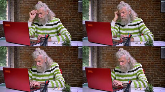 愉快的白人老人，浓密的白胡子，集中在电脑上打字，参与工作过程，砖砌办公室