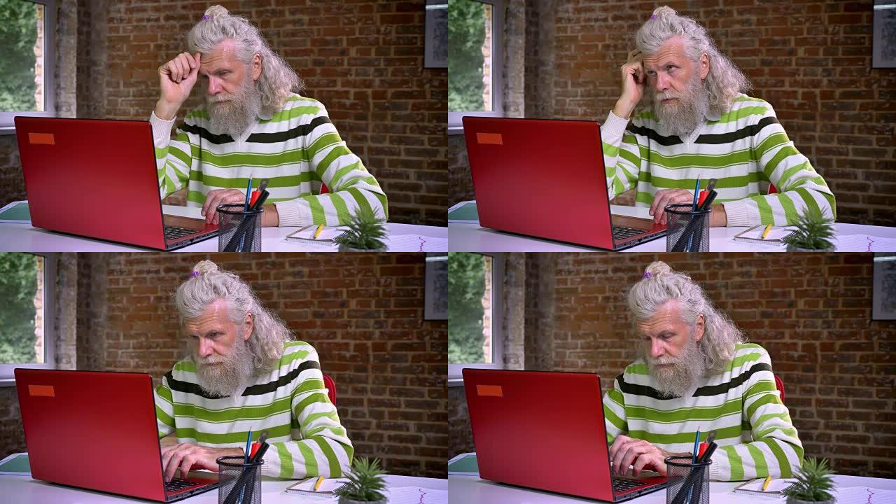 愉快的白人老人，浓密的白胡子，集中在电脑上打字，参与工作过程，砖砌办公室
