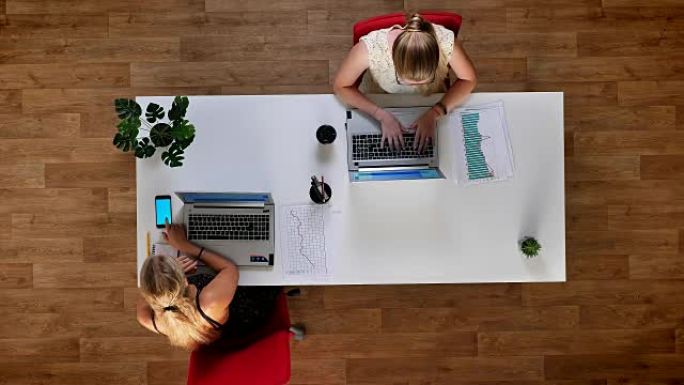 自上而下的镜头，两个富有成效的女孩坐在木制办公室的桌子旁，使用他们的笔记本电脑和蓝屏智能手机