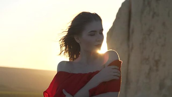 穿着红色连衣裙的年轻女子看着躲在悬崖后面的太阳风摇着她的头发
