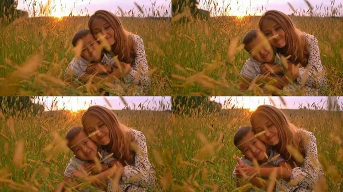 亚洲母亲温柔地抱着她的男孩，她在麦田的日落时露出拇指并快乐地微笑