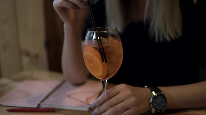 高兴的积极的金发女人在酒杯中享受鸡尾酒，用黑色吸管喝酒。戴着时尚的手表，她的日常组织者放在桌子上