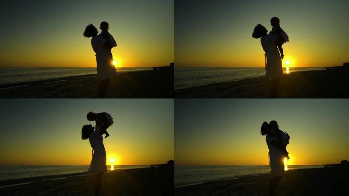 年轻漂亮的女人带着她的小女儿在日落时走在海边。妈妈把女儿抱在怀里。他们很高兴。夕阳映衬下的轮廓。慢动