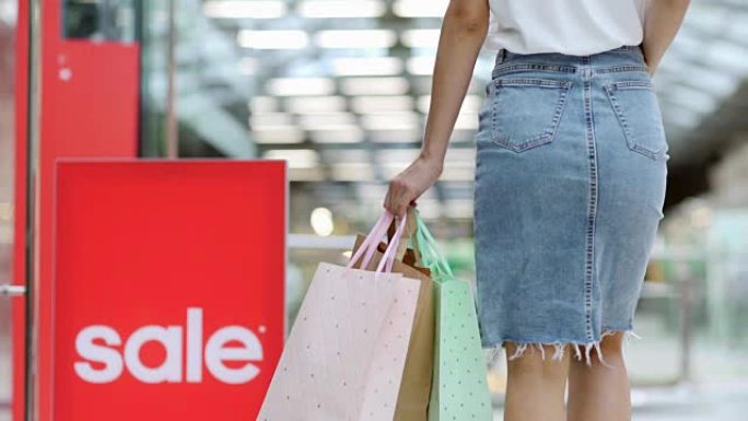 跟随穿着牛仔裙的面目全非的女人的镜头后视图，在销售当天慢动作在购物中心行走，用纸袋携带物品