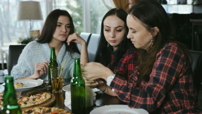 兴高采烈的年轻女孩在餐厅里看着智能手机上的照片。女孩吃披萨，喝啤酒，聊天，玩得开心。