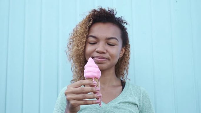 微笑的黑人女孩在蓝色墙壁背景下慢动作吃粉色融化冰淇淋