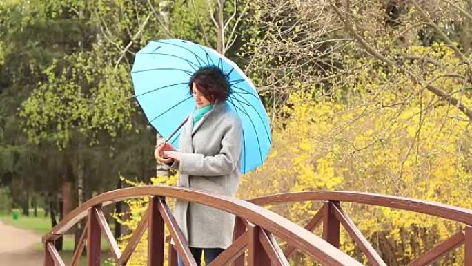 春天公园桥上拿着蓝色雨伞的年轻红头女人。绿树连翘开花背景。