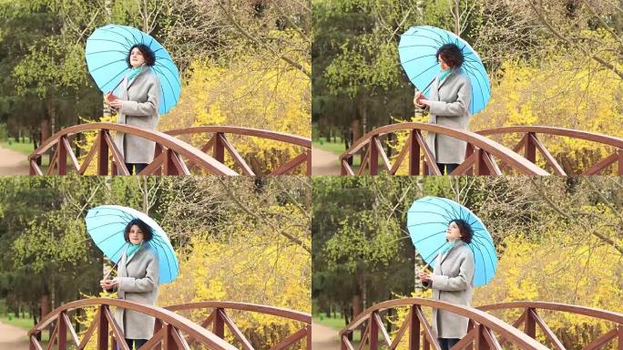 春天公园桥上拿着蓝色雨伞的年轻红头女人。绿树连翘开花背景。