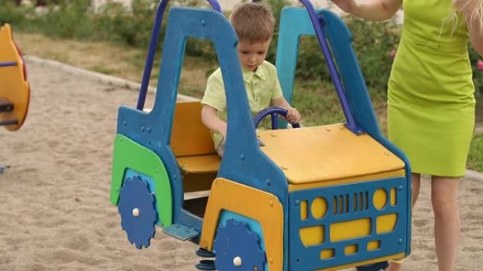 一个小男孩开着玩具车在操场上玩耍，他的母亲站在旁边