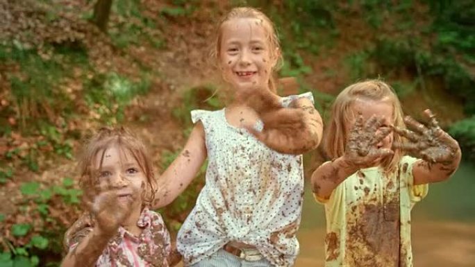 SLO MO站在森林小溪中，三个浑身是泥的女孩用泥泞的手向镜头挥手