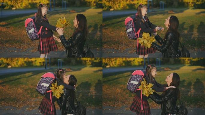 一个快乐的母亲在学校遇见了她的小女儿。妈妈用一束黄色的叶子拥抱和亲吻穿着校服的女儿。日落光线中的慢动