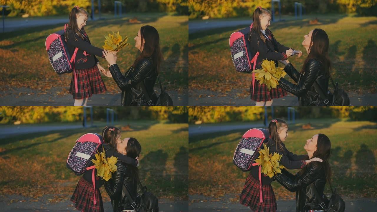 一个快乐的母亲在学校遇见了她的小女儿。妈妈用一束黄色的叶子拥抱和亲吻穿着校服的女儿。日落光线中的慢动