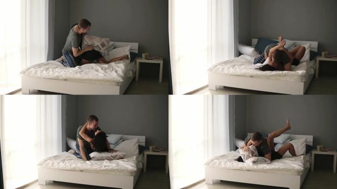 年轻的高加索夫妇在白色床单的床上用枕头打架，大笑，跌倒在床上并接吻。真爱，蜜月概念