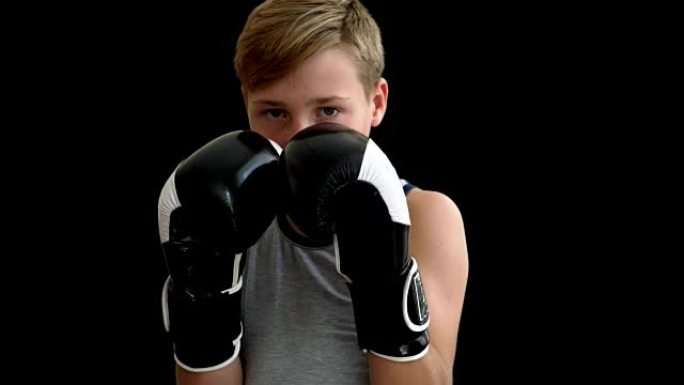 一名少年站在黑暗背景上的拳击手套中。这个男孩戴着黑白手套，几乎遮住了他的脸。这个男孩穿着灰色球衣，有