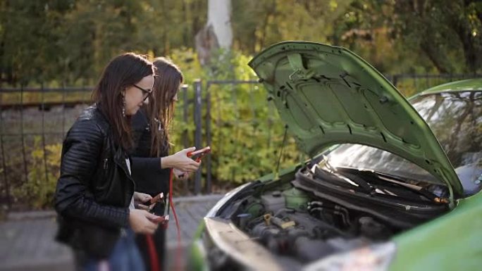 两个不太聪明的女孩试图给没电的汽车电池充电。两个女孩在汽车的敞篷上试图连接到电池引线进行充电