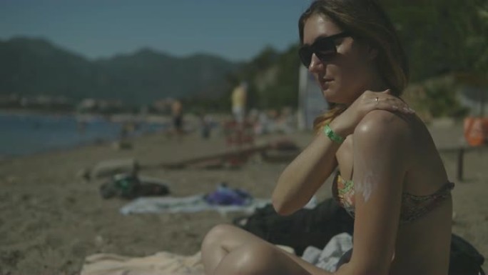 在阳光明媚的夏日，穿着泳衣的年轻女子在海滩上涂抹防晒霜。女孩在肩膀上涂抹防晒霜