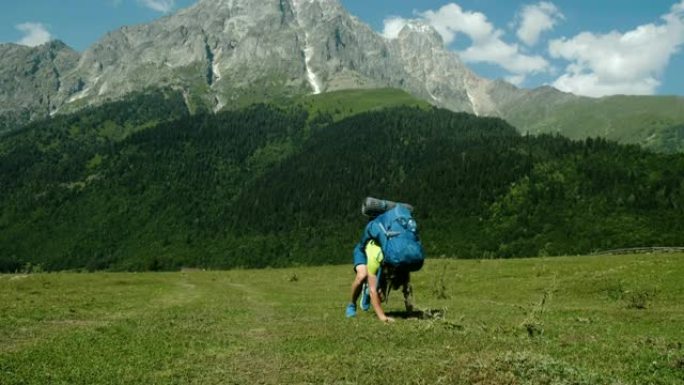 疲惫而虚弱的游客背着沉重的背包从地上爬起来，在美丽的山景背景下徒步旅行，一个病人慢慢站起来走路