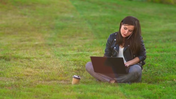 公园里草坪上的一个女孩在笔记本电脑上工作，电话铃响，接听来电。