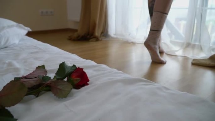 女性的腿在卧室明亮的窗户附近的脚趾上伸出来。红玫瑰在前景躺在床上。慢动作。