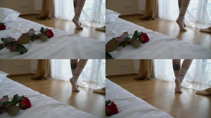 女性的腿在卧室明亮的窗户附近的脚趾上伸出来。红玫瑰在前景躺在床上。慢动作。