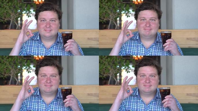 胖子在咖啡馆或餐馆里拿着一杯可乐饮料，对着镜头示意。