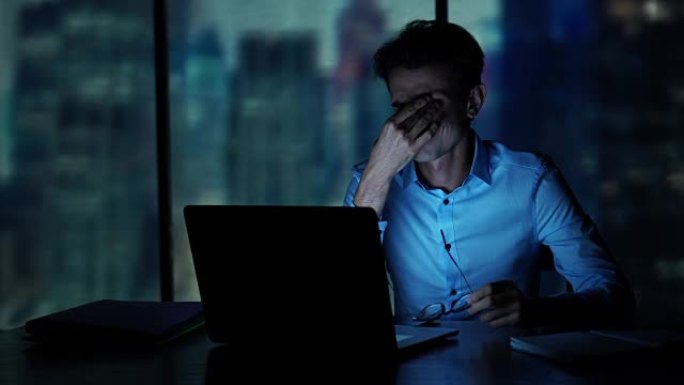 疲倦的年轻人深夜在办公室里用笔记本电脑工作。昏昏欲睡的商人坐在黑暗的办公室里
