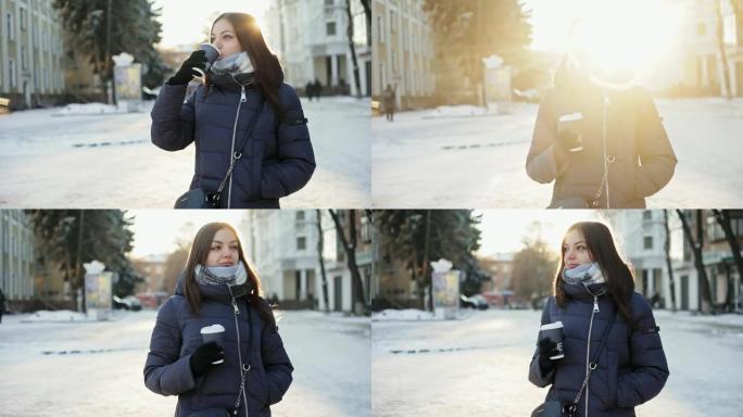 美丽的年轻女子在阳光明媚的冬天，在白雪皑皑的城市街道上，从阳光下闪耀着镜头，从纸杯里喝热咖啡